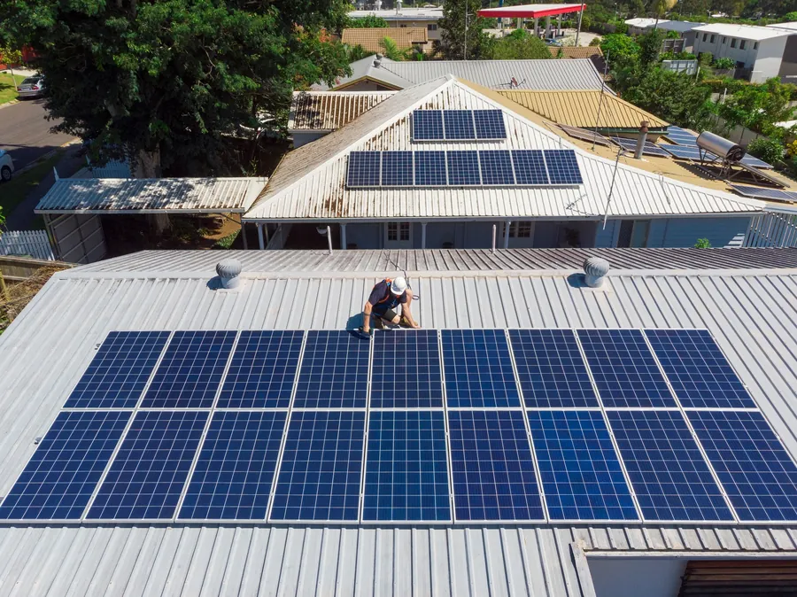An installer working on a rooftop solar array, with a roof-mounted solar hot water tank on the house behind